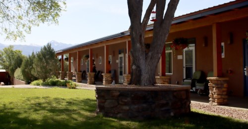 A row of rustic buildings with stone accents, surrounded by greenery and mountains in the background.