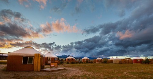 A scenic view of several yurts under a dramatic sky with clouds at sunset, surrounded by open land.