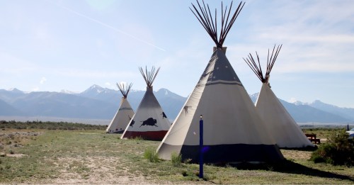 Four tipis stand in a grassy area with mountains in the background under a clear blue sky.
