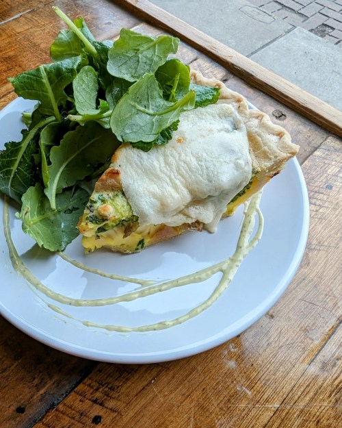 A slice of quiche topped with melted cheese, served with a side of fresh greens on a white plate.