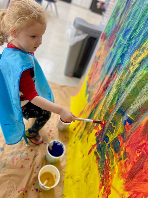 A young child in a blue smock paints a colorful mural on a large canvas, surrounded by paint containers.