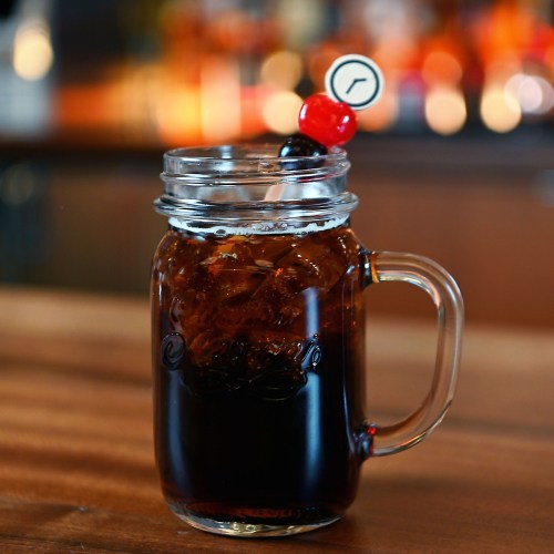 A mason jar filled with dark soda, ice, and a cherry garnish on a wooden bar.