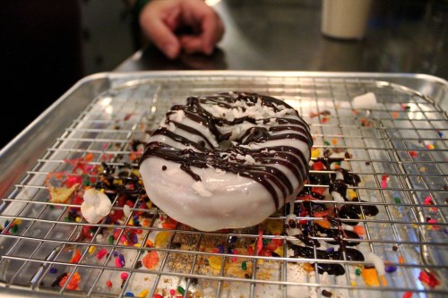 A frosted donut drizzled with chocolate and topped with sprinkles on a wire rack.