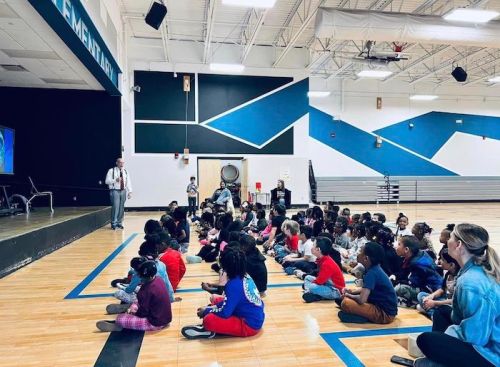 A group of children sitting on the floor in a gym, watching a presentation while an adult speaks.