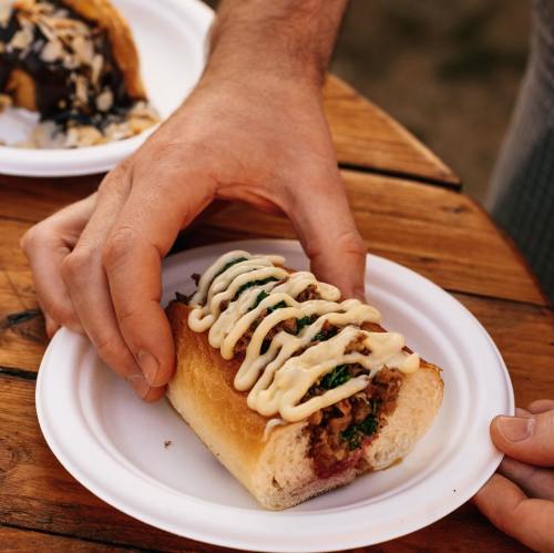 A hand holding a loaded sandwich with toppings, placed on a white paper plate on a wooden table.