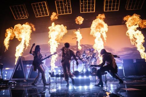 A dynamic rock band performs on stage with flames shooting up in the background, creating an energetic atmosphere.