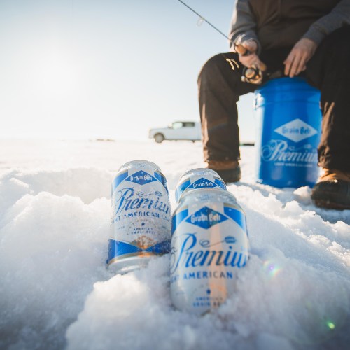 Cans of beer on snow with a person fishing in the background, a blue bucket and a truck visible in the distance.