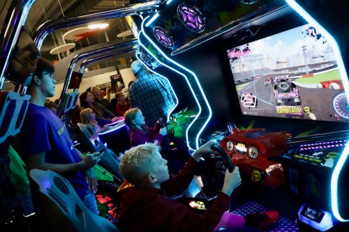 A child plays a racing arcade game while others watch in a lively arcade setting. Bright lights and colorful screens enhance the scene.