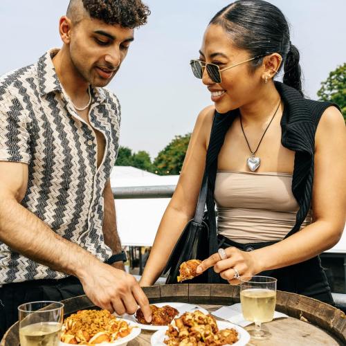 A man and woman enjoy food together at an outdoor event, smiling and sharing dishes with drinks in hand.
