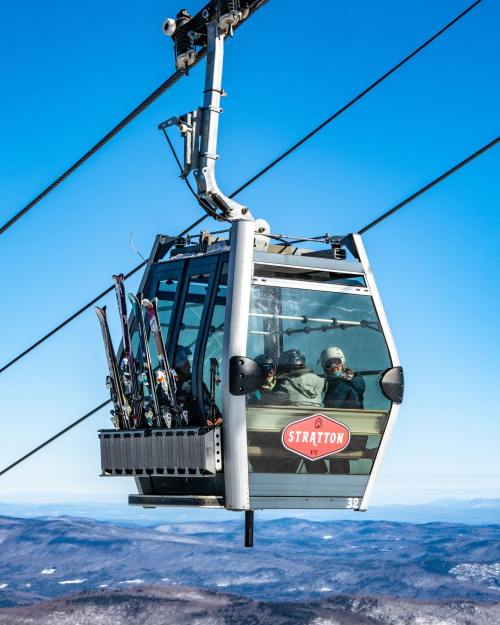 A ski gondola with passengers and skis ascends against a clear blue sky and mountain backdrop.