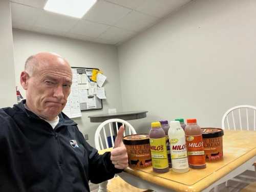 A man gives a thumbs up next to containers of Blue Bell ice cream and bottles of Milo's tea on a table.