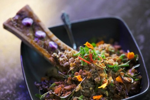 A black bowl filled with colorful rice, garnished with herbs and edible flowers, alongside a bone with toppings.