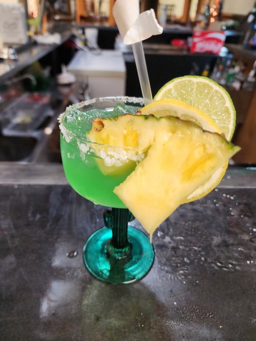 A vibrant green cocktail in a decorative glass, garnished with pineapple and lime slices, on a bar counter.