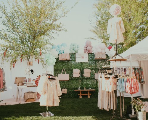 A vibrant outdoor market stall featuring clothing, bags, and colorful balloons against a green backdrop.