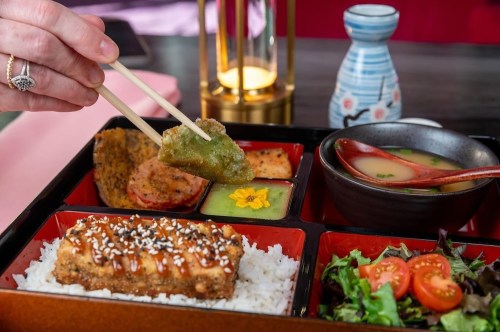 A hand using chopsticks to pick up food from a colorful bento box with rice, salad, and soup.
