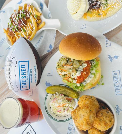 A spread of food featuring a sandwich, pickles, coleslaw, fried biscuits, and a football, with a beer in the background.