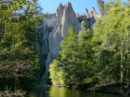 Tall rock formations rise above a serene river, surrounded by lush green trees under a clear blue sky.
