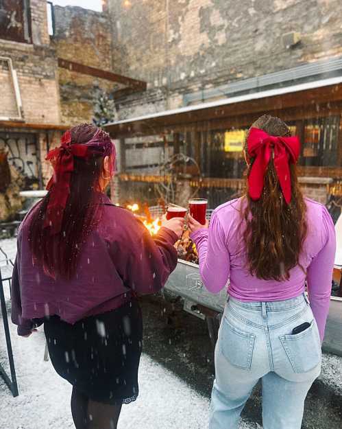 Two women with red bows in their hair hold drinks, standing by a fire in a snowy outdoor setting.