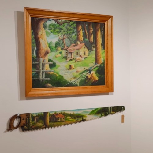 A framed landscape painting of a cabin in the woods, with a decorative saw mounted below it.