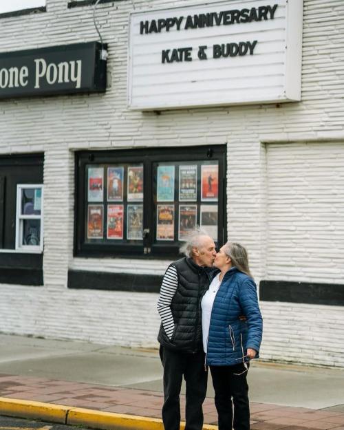 An elderly couple kisses in front of a venue celebrating their anniversary with a sign that reads "Happy Anniversary Kate & Buddy."