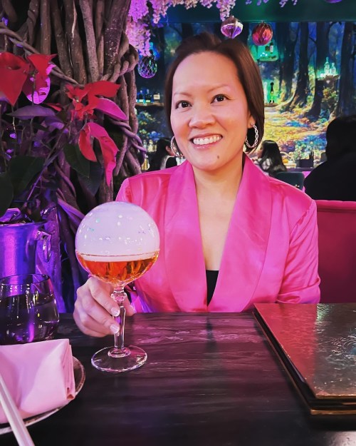 A woman in a pink blazer smiles while holding a cocktail with a frothy top, seated in a colorful, vibrant setting.