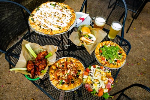 A table with various pizzas, wings, a salad, and drinks, set outdoors on a black metal table.