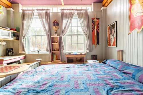 Cozy bedroom with a colorful quilt, large windows, and decorative banners on the walls. Bright and inviting atmosphere.