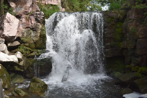 A cascading waterfall flows over rocky terrain, surrounded by lush greenery and moss-covered stones.