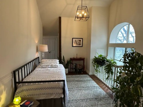 Cozy bedroom with a bed, lamp, plants, and large window, featuring warm lighting and a welcoming atmosphere.