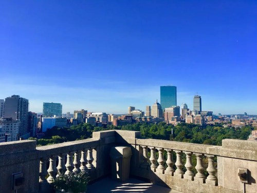 A panoramic view of a city skyline with tall buildings and clear blue skies, framed by a stone balcony.