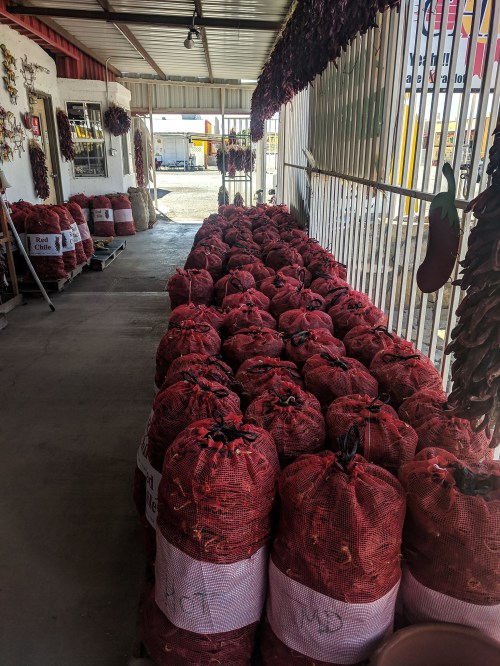 Bags of red chili peppers lined up in a market, with a colorful display of dried peppers in the background.