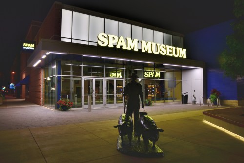 Exterior of the Spam Museum at night, featuring a large sign and a statue of a person with pigs in front.
