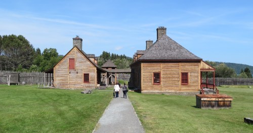 Two people walk along a path toward two historic wooden buildings in a grassy area under a clear blue sky.