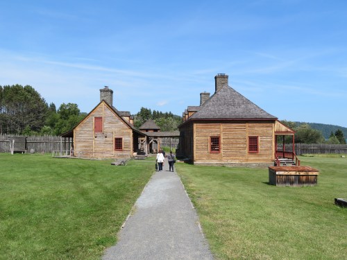 Two people walk down a path toward historic wooden buildings surrounded by green grass and trees under a blue sky.