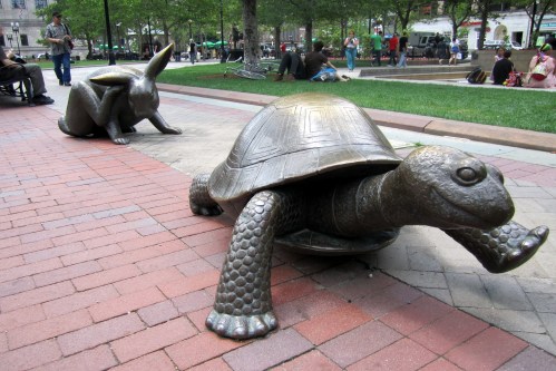 Bronze sculptures of a turtle and a rabbit in a park, with people sitting and walking in the background.
