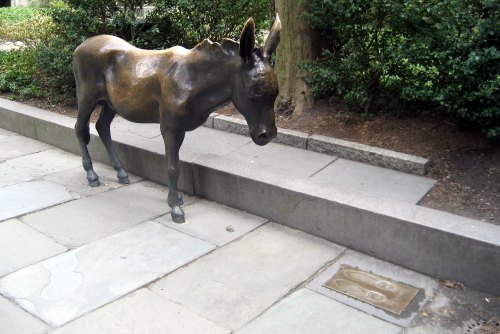 A bronze statue of a donkey stands beside a plaque on a stone pathway, surrounded by greenery.