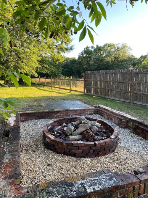 A circular brick fire pit surrounded by gravel, set in a grassy area with trees and a wooden fence in the background.