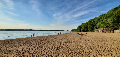 A sandy beach by a calm lake, with people swimming and relaxing under a clear blue sky and green trees in the background.