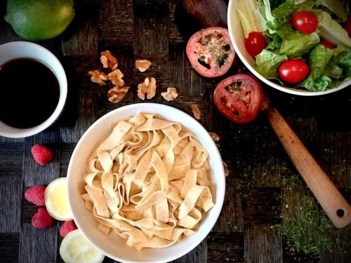 A bowl of pasta surrounded by fresh vegetables, nuts, and a small cup of sauce on a textured surface.