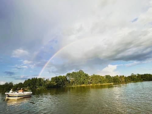 A boat on a calm river with a rainbow arching over green trees and a partly cloudy sky.
