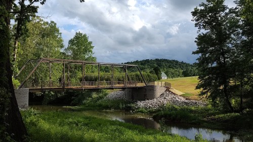 A rustic metal bridge spans a calm stream, surrounded by lush greenery and rolling hills under a cloudy sky.