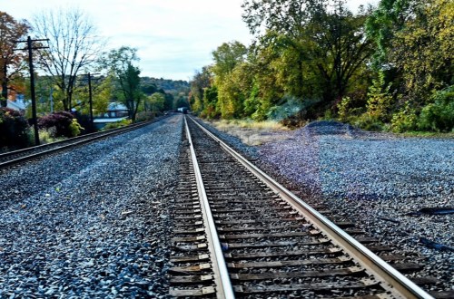 A view of railway tracks stretching into the distance, flanked by trees and gravel under a clear sky.