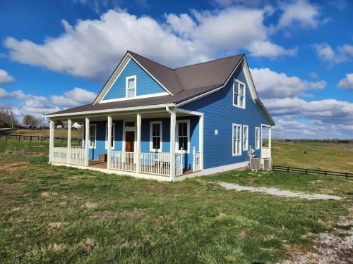 A blue farmhouse with a porch, surrounded by green grass and a cloudy sky.