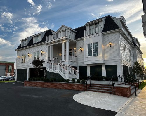 A large, elegant white house with a porch, surrounded by greenery and a paved driveway, under a blue sky.