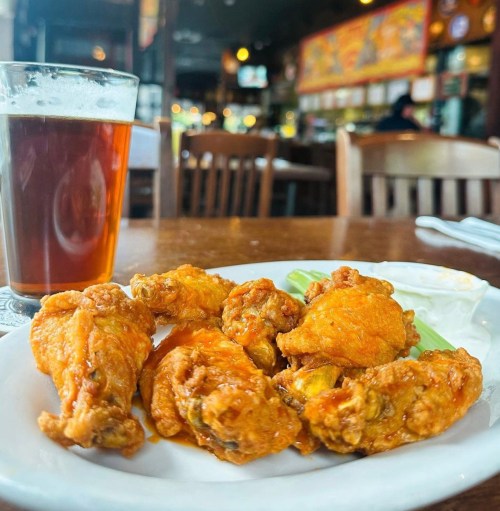 A plate of crispy chicken wings with celery and dipping sauce, alongside a glass of amber beer in a cozy restaurant.
