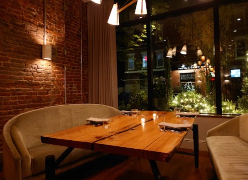 Cozy restaurant interior with a wooden table, soft seating, and a view of outdoor lights through large windows.
