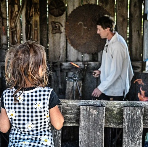 A child watches a blacksmith at work in a rustic workshop, surrounded by tools and wooden walls.