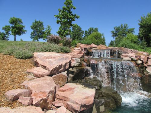 A serene waterfall cascades over pink rocks, surrounded by lush greenery and blue skies.