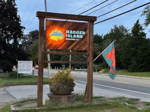 Sign for Ragged Island Brewing Co. with a glowing logo, surrounded by greenery and a flag, along a roadside.
