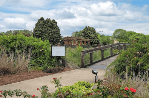 A wooden bridge leads through lush greenery to a historic building under a cloudy sky.